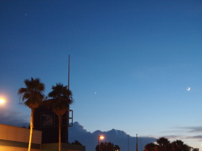 [The upper portions of the sky are becoming a darker blue, but the rest is still a lighter blue. There are several bright lights near the trees and building and some dark clouds at the horizon, but the rest of the sky is clear except for the three orbs, Jupiter, Venus, and the moon, already described.]